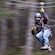 A bearded man wearing a helmet, harness and gloves smiles as he zooms through the forest on a zip line